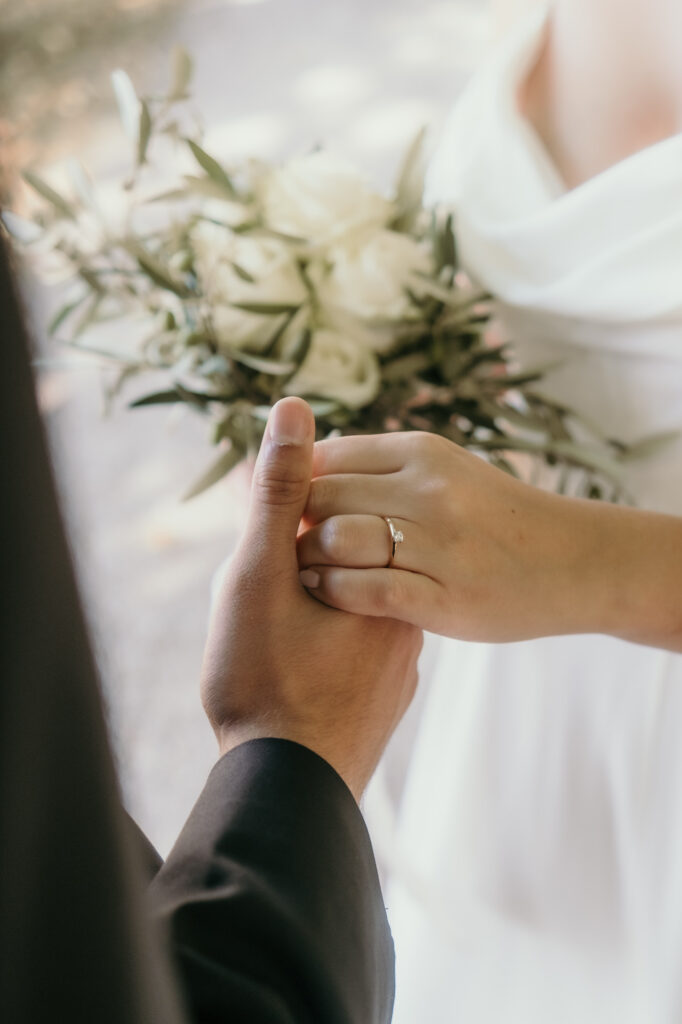 Diese Nahaufnahme zeigt die verschränkten Hände des Hochzeitspaares, als sie voreinander stehen.