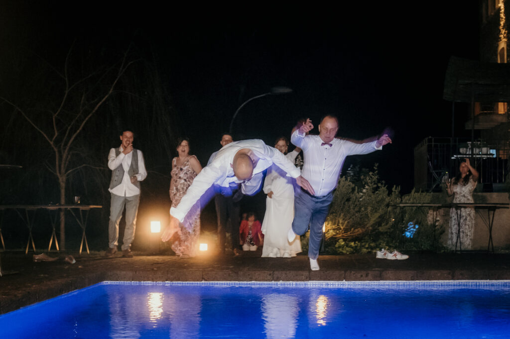 Zwei männliche Gäste springen kopfüber in den Pool. Dahinter jubelt das Hochzeitspaar.