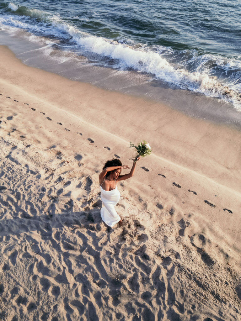 Ein Foto aus der Luft betrachtet sieht man ein Brautmodel mit ihrem Strauß am Meer tanzen.