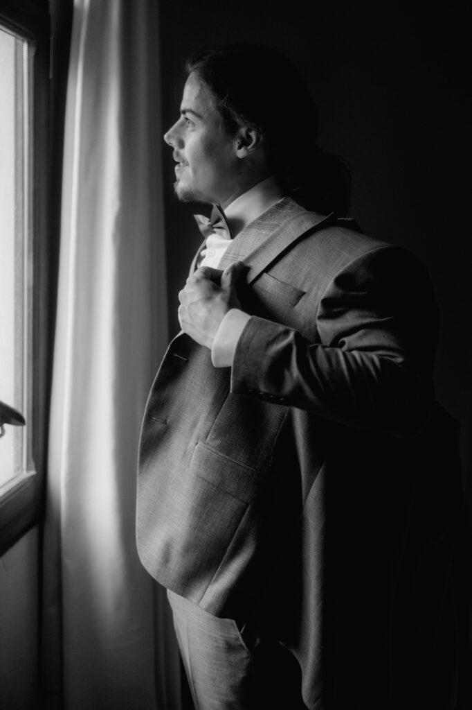 Der Bräutigam steht vor einem Fenster, während er sich sein Sakko anzieht und sieht hinaus.