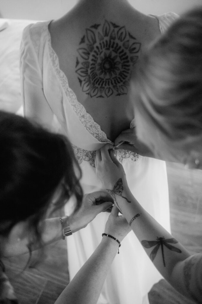 Das Brautkleid wird von zwei Damen geschlossen. Das große Rücken Tattoo der Braut sticht heraus.
