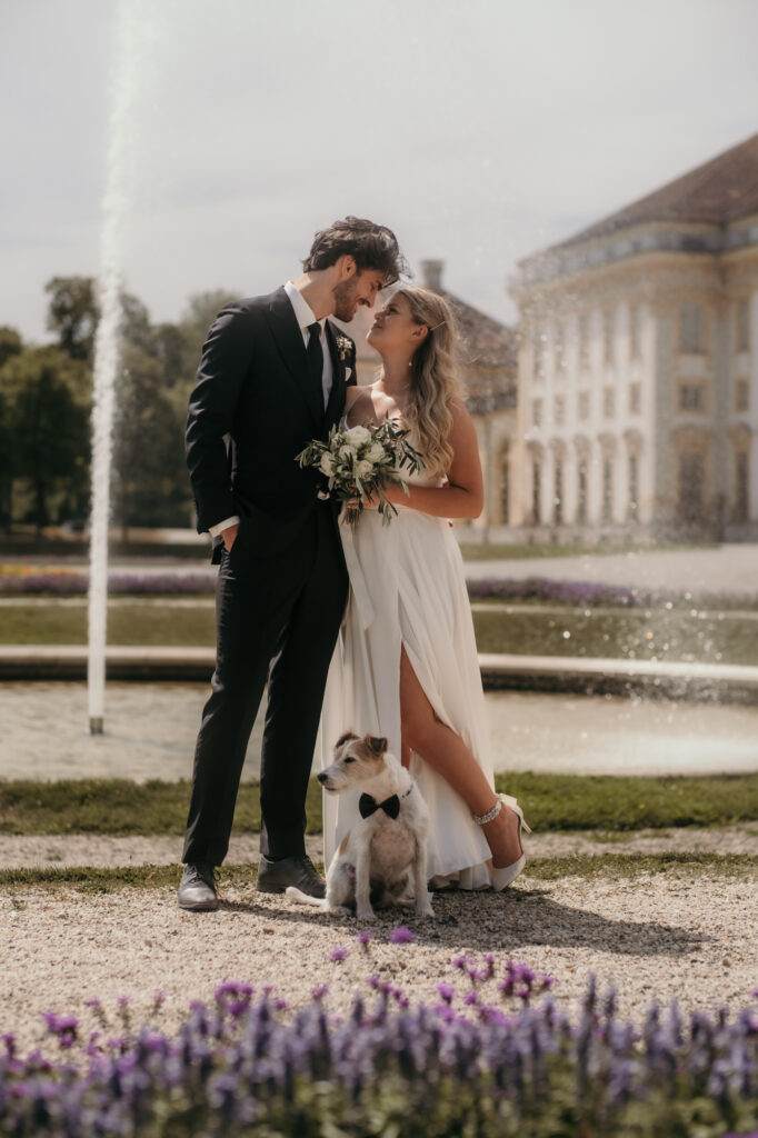 Dieses Hochzeitsfoto zeigt das Paar mit ihrem Hund vor dem Brunnen des Schlosses Schleißheim.