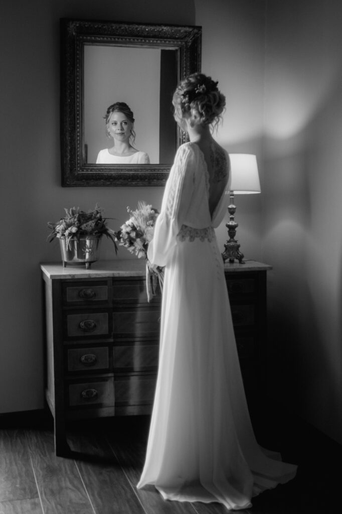 Sich in einem Wandspiegel betrachtend steht die Braut vor einer historischen Kommode.