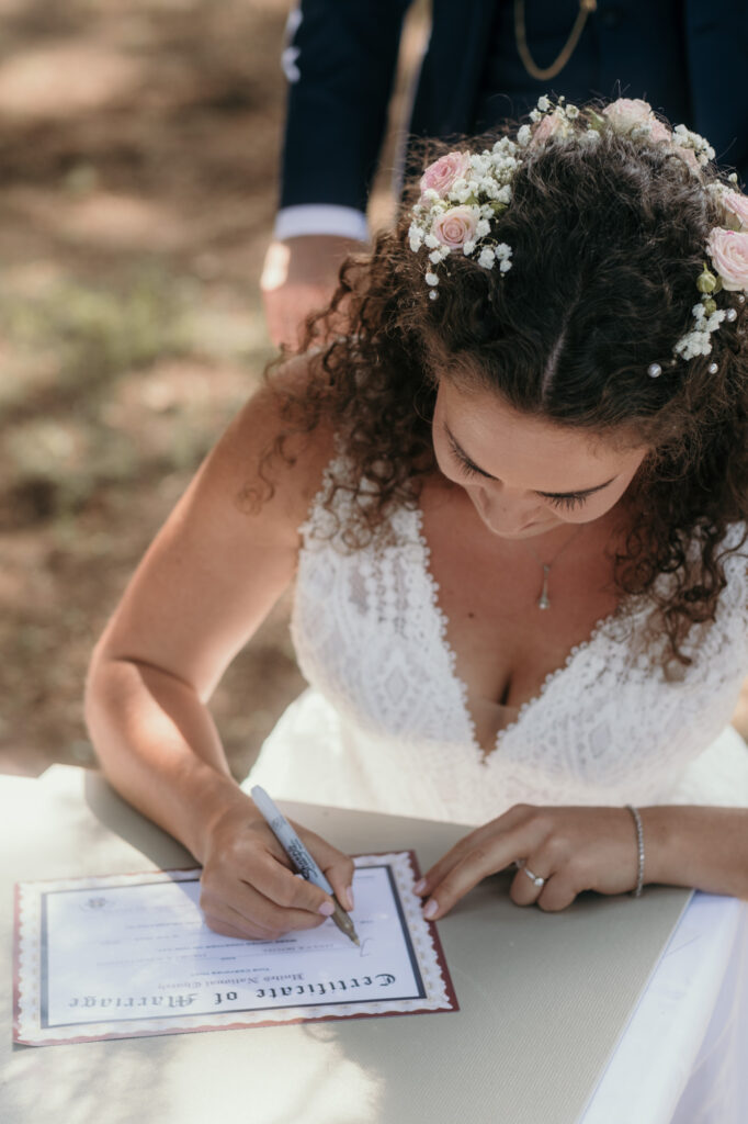 Die Braut unterschreibt an einem Tisch sitzend die Hochzeitsurkunde. Der Bräutigam steht hinter ihr.