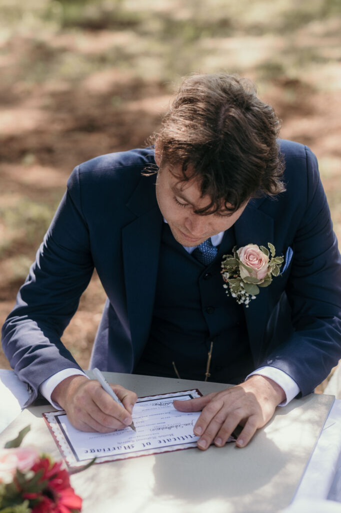 Der Bräutigam unterschreibt die Hochzeitsurkunde an einem Tisch sitzend.