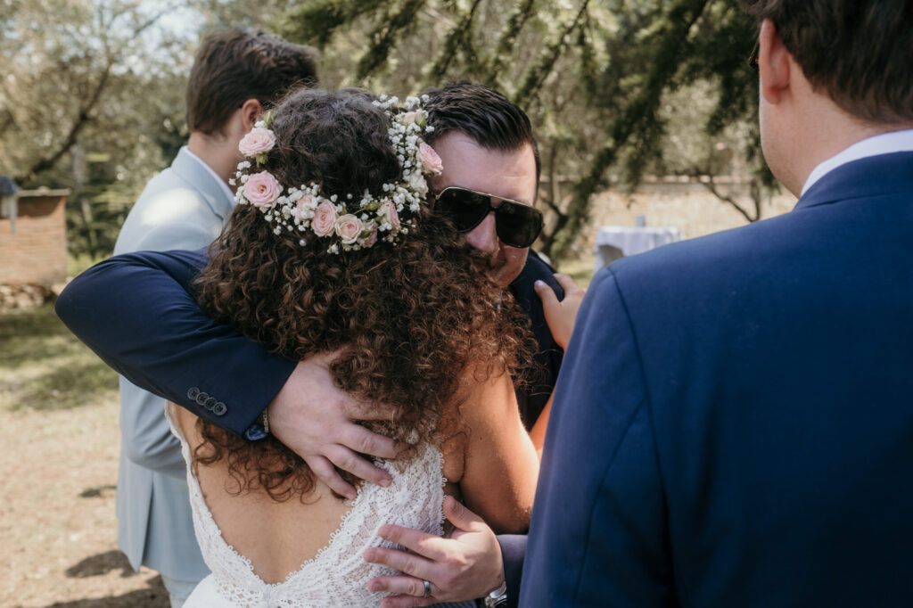 Die Braut wird überschwänglich von einem Herren mit Sonnenbrille umarmt.