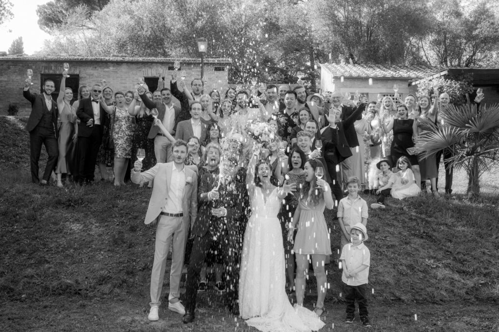 Auf diesem Gruppenfoto der ganzen Hochzeitsgesellschaft spritzt der Bräutigam mit einer Champagnerflasche während alle jubeln.