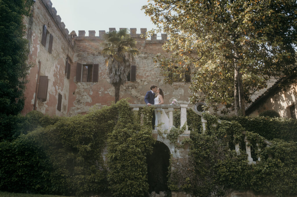 Auf einer Balustrade des historischen Herrenhauses steht das Hochzeitspaar von fern betrachtet und küsst sich. 