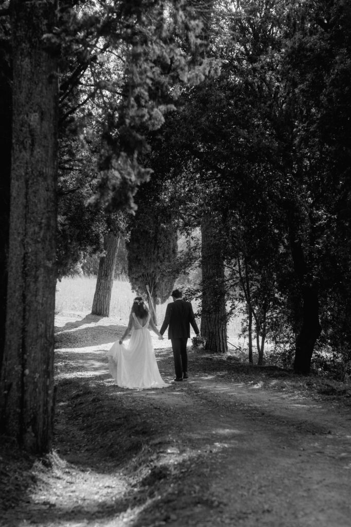 Durch eine Allee spaziert das Hochzeitspaar auf einem Feldweg Händchen haltend von der Kamera weg.