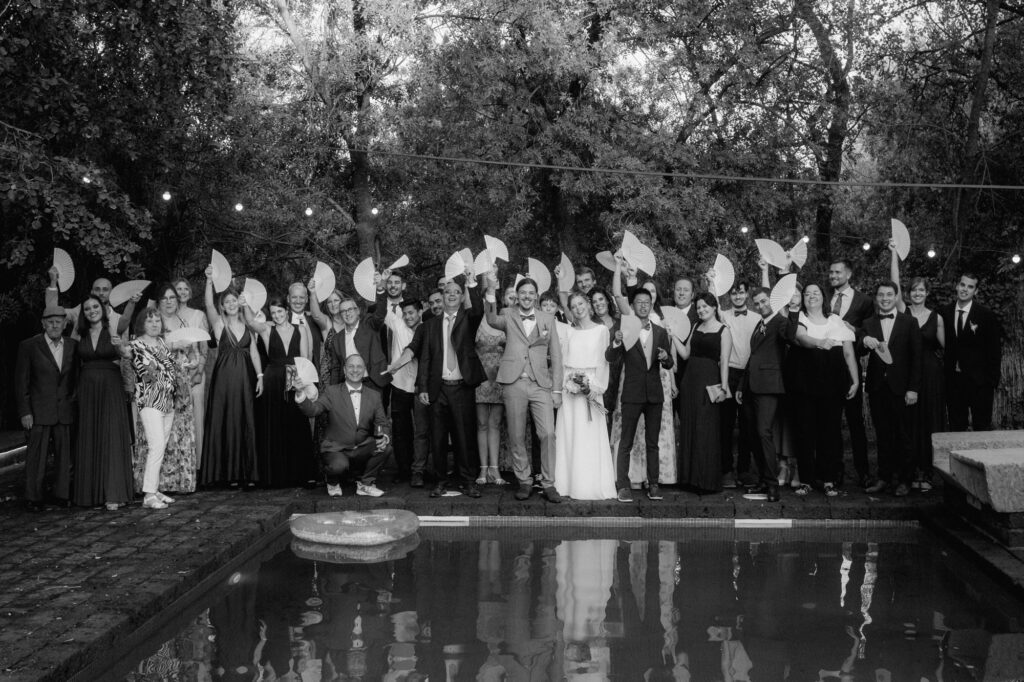 Dieses Gruppenfoto zeigt die ganze Hochzeitsgesellschaft vor einem Pool. Alle halten weiße Fächer hoch.