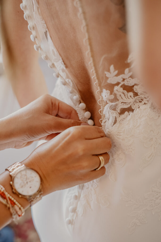 Zwei Damenhände helfen der Braut beim Schließen der langen Knopfreihe an ihrem Brautkleids.