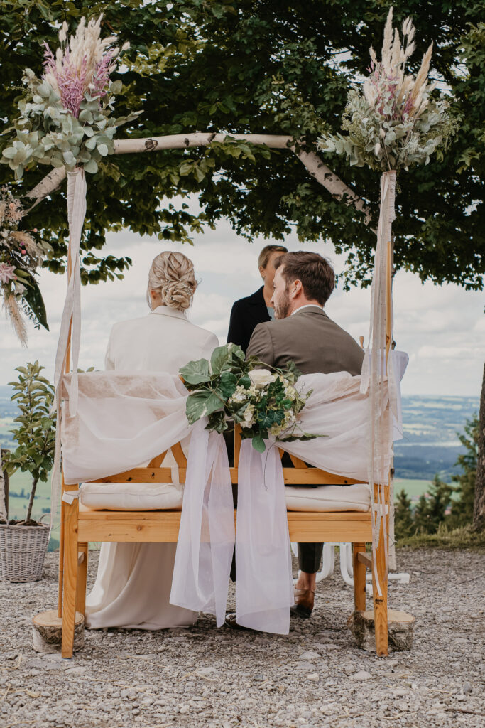 Das Hochzeitspaar sitzt von hinten betrachtet auf der festlich mit Blumen geschmückten Bank.