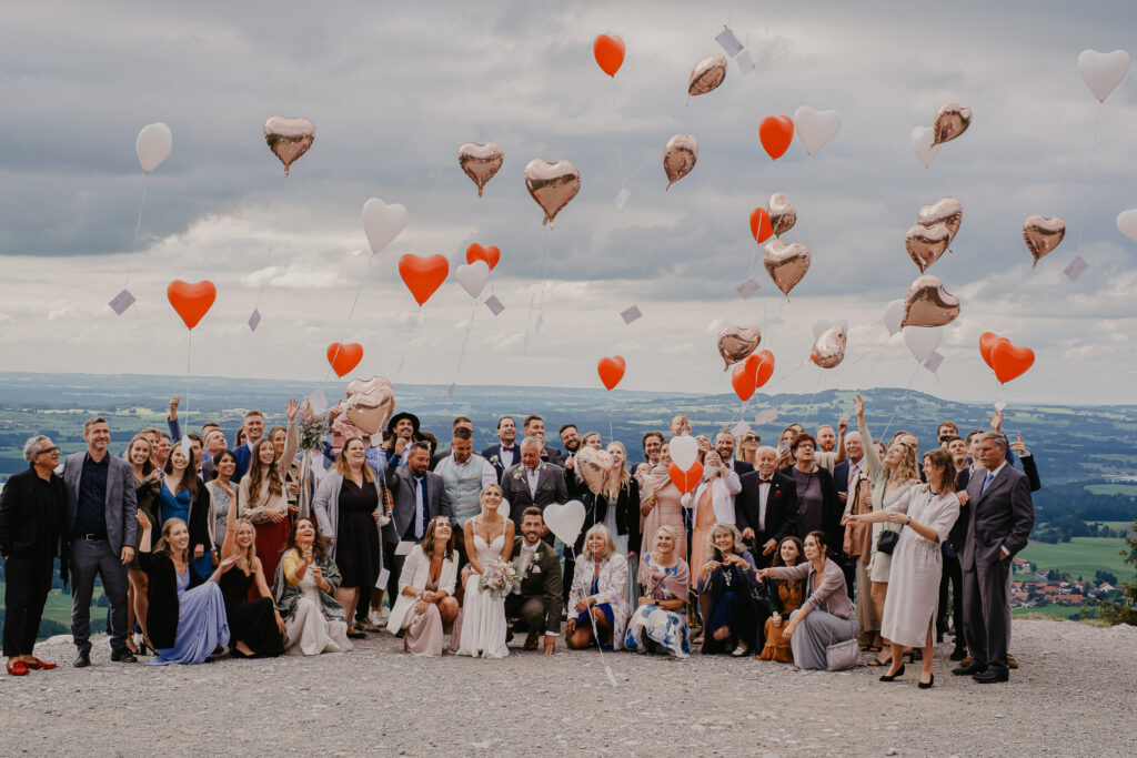 Vor der Landschaft der bayerischen Alpen ist die Hochzeitsgesellschaft für ein Gruppenfoto versammelt und lässt Herzluftballons in die Luft steigen.