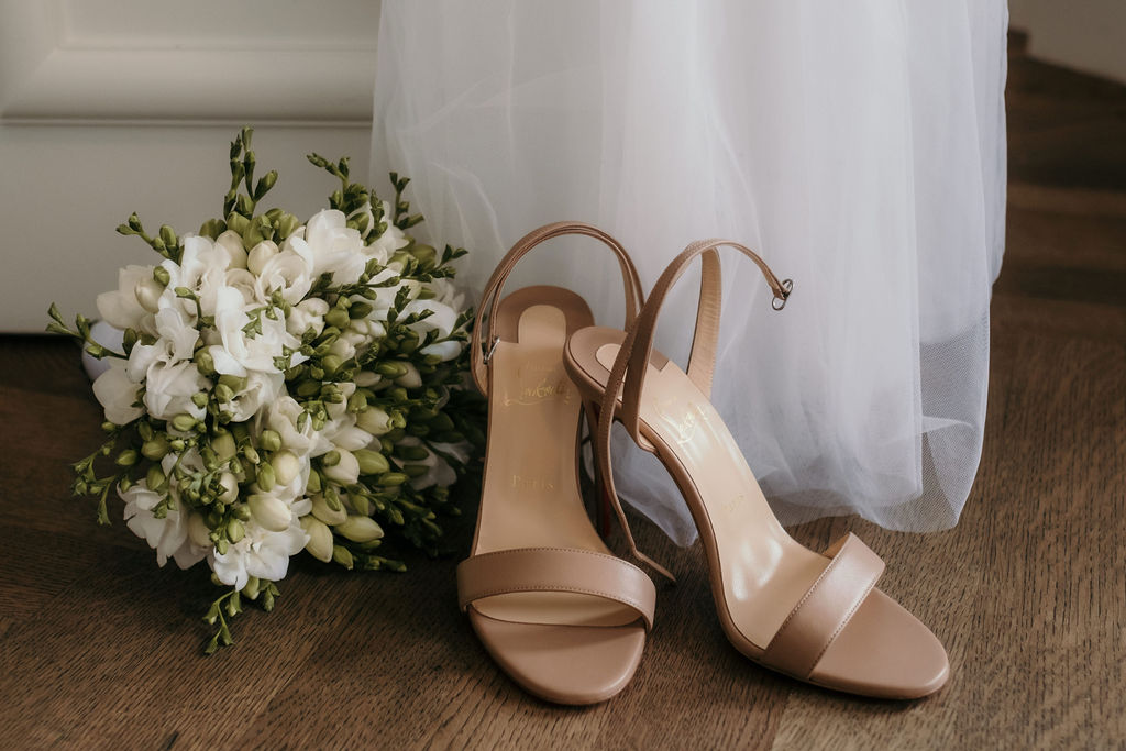 Ein Brautstrauß und High Heels stehen auf dem Boden vor einem Hochzeitskleid