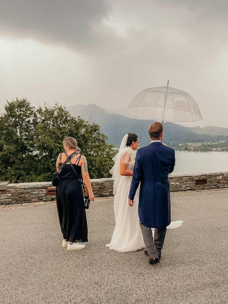 Beim Hochzeitspaarshooting im Regen hat das Paar einen durchsichtigen Regenschirm, Karin steht daneben.