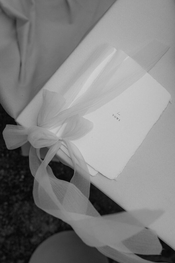 A black and white photo of two vow cards laying on a table with the wind blowing through the ribbons
