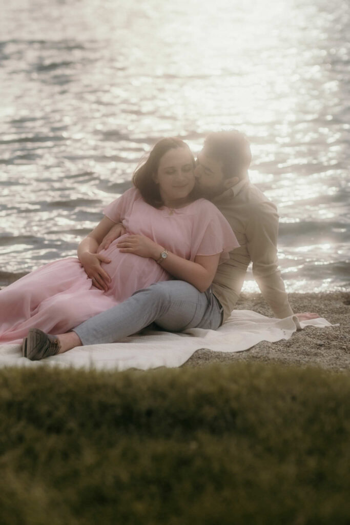 A pregnant couple sitting on the ground in front of a lake cuddling and smiling. 