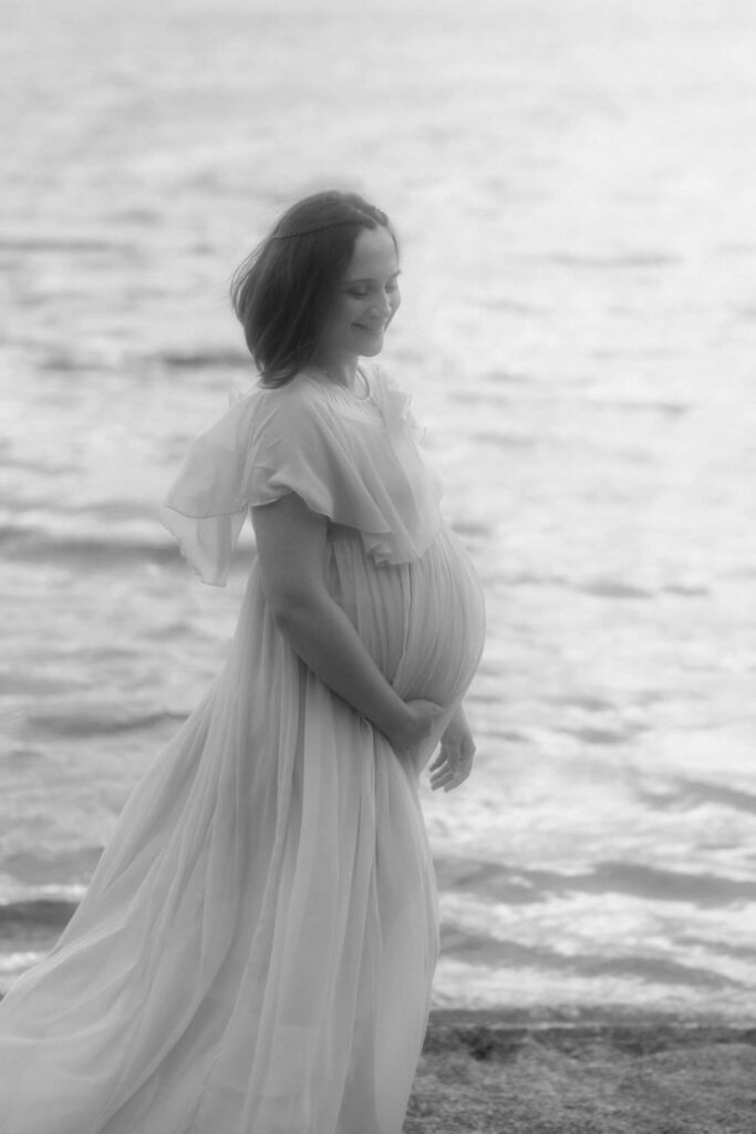Pregnant woman standing in front of a lake smiling down at her baby belly.