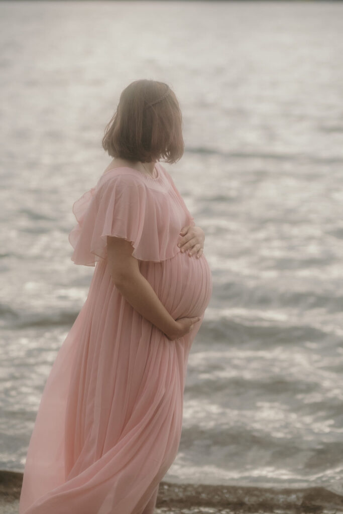 Pregnant woman standing in front of a lake holding her baby belly and looking off into the distance.