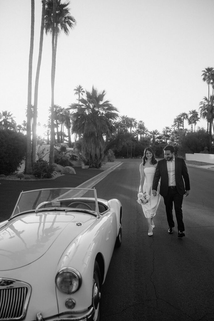 A couple walking towards their wedding car holding hands with palm trees in the background