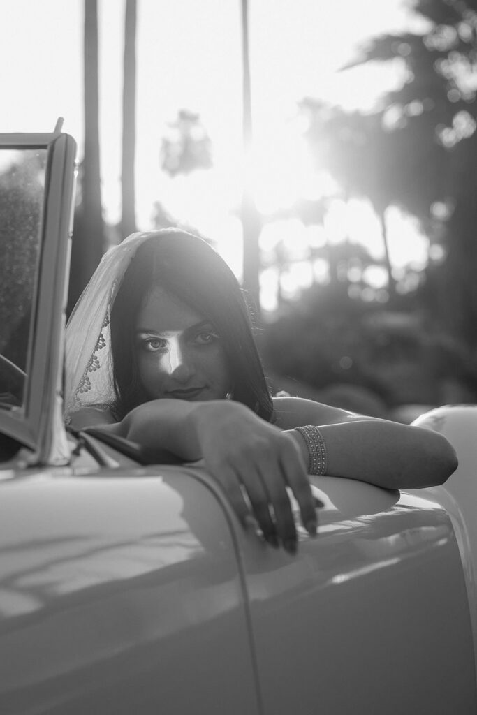 A bride sitting in a convertible leaning over the door looking directly into the camera