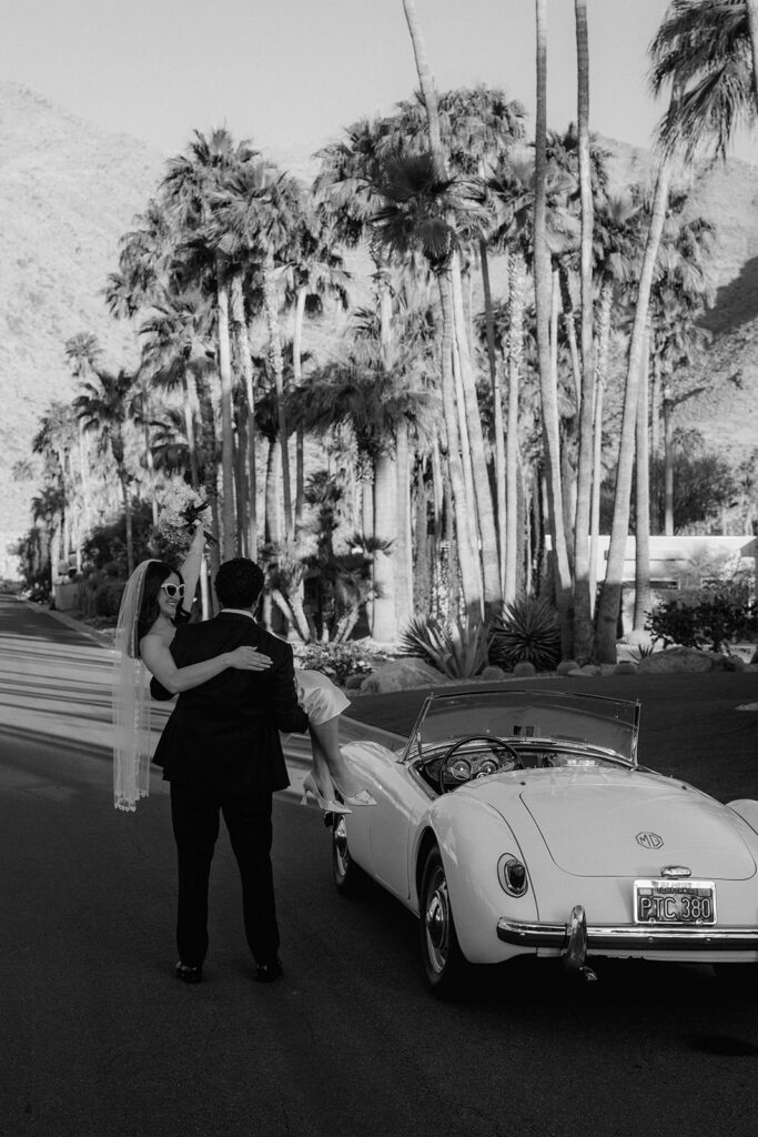 The groom carrying the bride towards their wedding car with a palm tree lined street in the background
